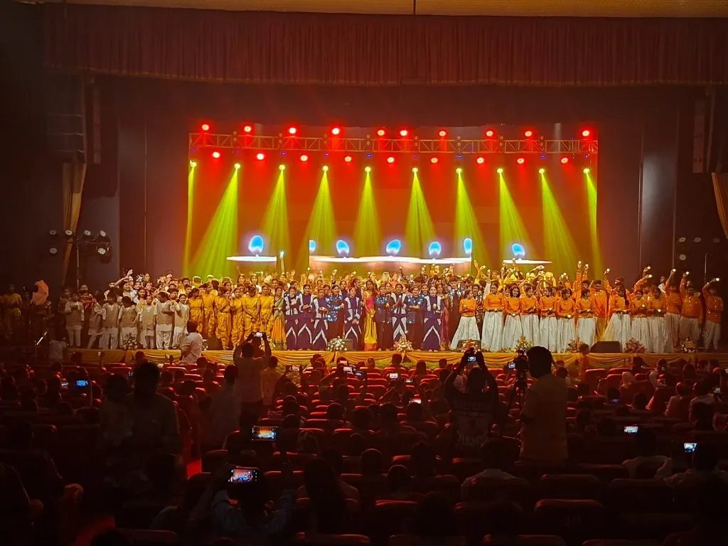 Large group of performers in coordinated costumes on a lit stage during a cultural production, audience capturing the moment in the foreground